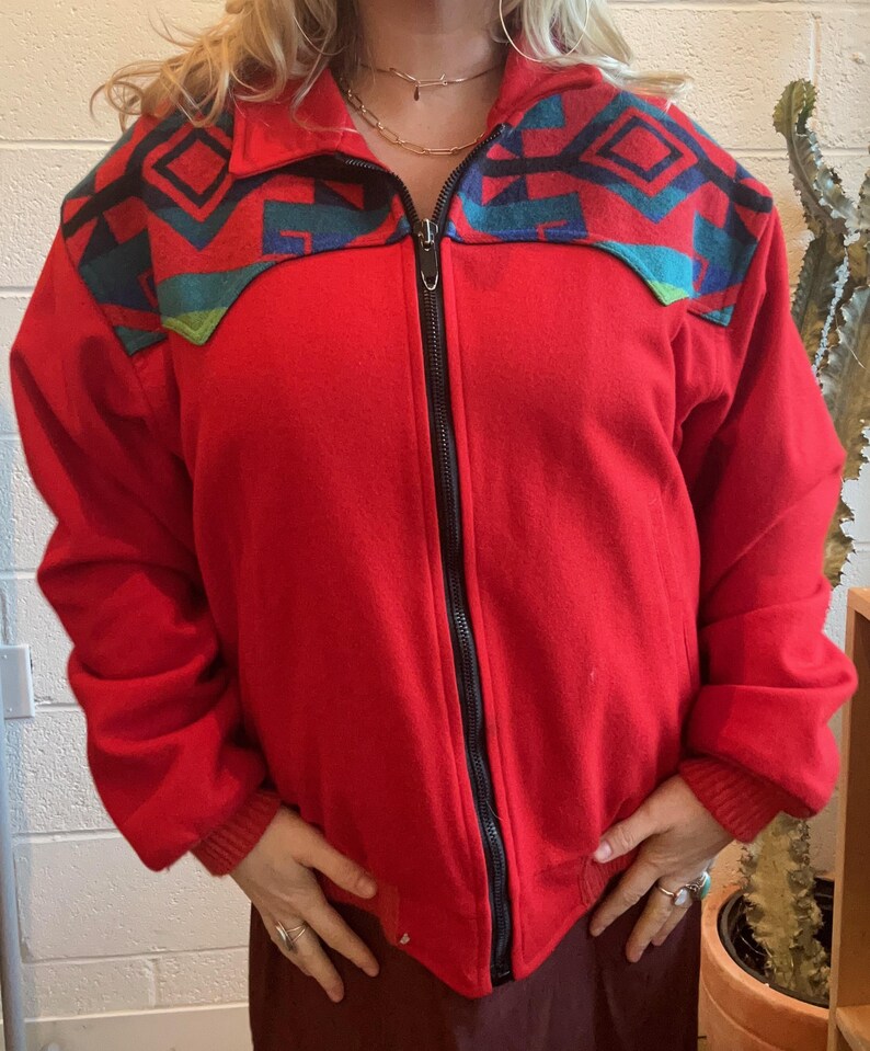 May include: A red zip-up jacket with a geometric pattern in teal, black, and turquoise across the shoulders. The jacket has a black zipper and ribbed cuffs. The jacket is a solid red color.
