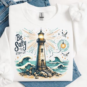 以下が含まれることがあります： 白いスウェットシャツに、灯台と「Be Salty Stay Lit」の文字がプリントされています。灯台は波と星に囲まれています。