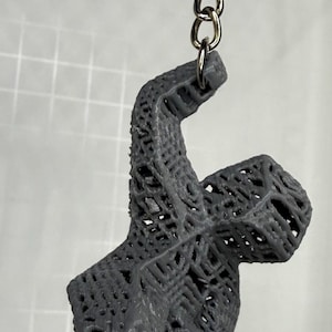 May include: A gray, 3D-printed keychain featuring a stylized figure with an open, lattice-like design. The figure is attached to a silver chain and keyring. The keychain is made of plastic.