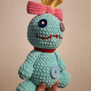 May include: A light blue crocheted plush doll with a textured yarn surface. It features a pink bow, yellow tufted hair, a red collar, and mismatched button eyes in dark blue and purple. A red stitched mouth and a large light blue button on the belly.