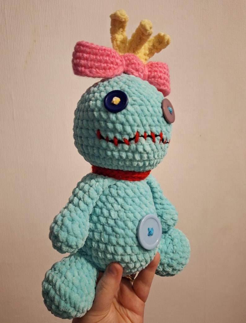 May include: A light blue crocheted plush doll with a textured yarn surface. It features a pink bow, yellow tufted hair, a red collar, and mismatched button eyes in dark blue and purple. A red stitched mouth and a large light blue button on the belly.