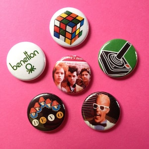 Puede incluir: Siete botones coloridos con diferentes diseños. Los botones muestran un cubo de Rubik, un joystick, una banda llamada DEVO, una marca de moda llamada Benetton y un grupo de personas.