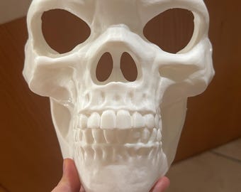May include: A white skull mask with large eye sockets, a prominent nose cavity, and a full set of teeth. The mask is made of a solid material and is designed to be worn. The mask is a Halloween costume.