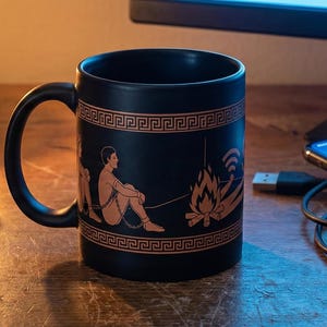 May include: A black ceramic mug with a Greek-inspired design featuring a seated figure and a fire. A copy of Plato's "The Republic" and a lit candle are nearby. A smartphone is charging on the desk.