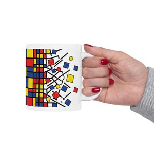 May include: White ceramic mug featuring a colorful geometric design with red, yellow, and blue squares and black lines. The mug has a white handle and is held by a person with red nail polish.