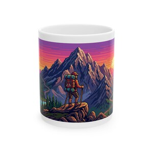 May include: White ceramic mug featuring a pixelated illustration of a hiker with a backpack and trekking poles, standing on a rocky outcrop. The background depicts a mountain range under a sunset sky with shades of orange, purple, and yellow.
