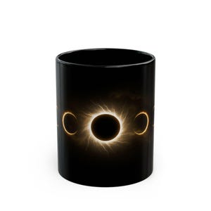 May include: A black ceramic mug featuring a graphic print of a solar eclipse. The design displays a central dark circle surrounded by a glowing golden corona with radiating light streaks, flanked by two crescent moon phases on either side.