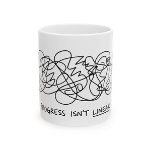 May include: A white ceramic mug featuring a black ink-style scribble design wrapping around the center. Below the abstract line art, the text "PROGRESS ISN'T LINEAR" is printed in a simple, black, hand-drawn style font with a line under the word LINEAR.