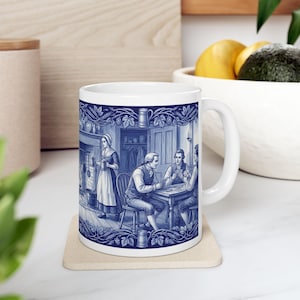 May include: White ceramic mug with a blue and white illustration depicting a historical scene of people playing cards and a woman serving drinks. The mug has a curved handle and a decorative border of vines and leaves.