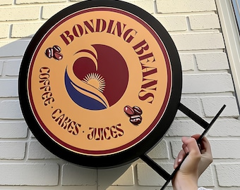 May include: A circular black-framed sign with a tan background featuring the text BONDING BEANS, COFFEE, CAKES, JUICES. The center graphic displays a stylized sun rising over a blue wave within a maroon circle, accompanied by two coffee bean icons.