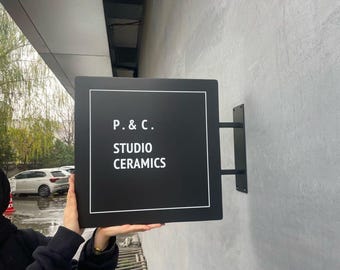 May include: A black square sign with white text that reads "P. & C. STUDIO CERAMICS." The sign is mounted on a gray wall with a black metal bracket. The sign is held by a person.