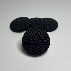 May include: A collection of five black, circular objects with a textured surface and the word "HAXER" embossed on each. The objects are arranged on a white background, with one in the foreground and the others slightly behind.