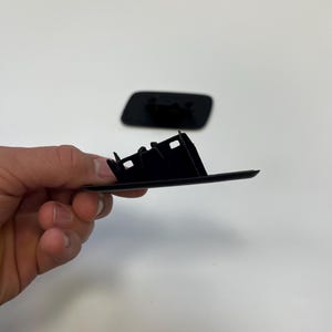 Può includere: Un componente automobilistico in plastica nera con una base rettangolare e diverse sezioni angolate rialzate. Il componente è tenuto su uno sfondo bianco, con un secondo componente simile visibile sullo sfondo.