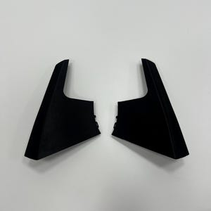 May include: Two black, angular automotive interior trim pieces. Each piece has a triangular shape with a flat top and a textured surface. The pieces are symmetrical and appear to be designed for the left and right sides of a vehicle.