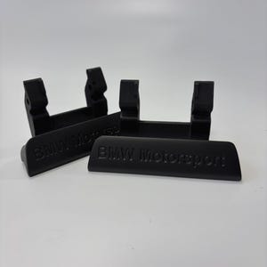 May include: Four black automotive parts with the text "BMW Motorsport" embossed on them. The parts have a rectangular base with two vertical supports. The parts are made of a matte black material.