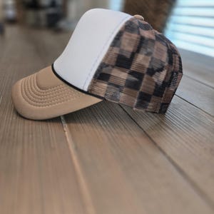 May include: A trucker hat featuring a solid white front panel, a tan curved brim, and a mesh back panel with a brown and black checkered pattern. The hat includes a thin black piping detail separating the front panel from the side mesh section.