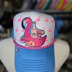May include: A blue and white trucker hat with a pink flamingo design. The flamingo is floating on a pool float with a bottle and a cocktail glass. The hat has a pink chain and a small charm.