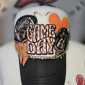 May include: A white and black baseball cap with "GAME DAY" in orange and white checkered letters. The cap features a black brim, gold chain, and patches of a football, orange heart, and a black and gold design.