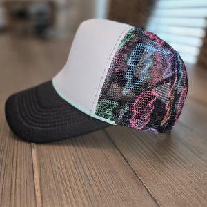 May include: A trucker hat featuring a solid black brim, a light gray front panel, and a black mesh back panel. The mesh back is decorated with a vibrant, multi-colored lightning bolt pattern in shades of pink, blue, and green, accented by a thin light blue trim.