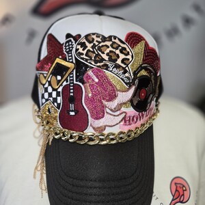 May include: A white and black trucker hat featuring various embroidered patches including a leopard print cowboy hat, pink glittery boots, a guitar, a vinyl record, and a musical note. The hat is accented with a gold chain across the brim.