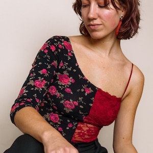 May include: A woman wearing a black and red floral top with a one-shoulder design. The top has a lace panel on the red side. She is wearing black trousers and has short, wavy brown hair.
