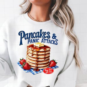 Könnte beinhalten: Weißer Pullover mit einem Grafikdruck einer Stapel Pancakes mit Sirup und Butter und dem Text "Pancakes & Panic Attacks" in Blau. Die Grafik enthält auch Erdbeeren und eine blau-weiß karierte Tischdecke. Der Text "Buy One Get One" ist in Rot.