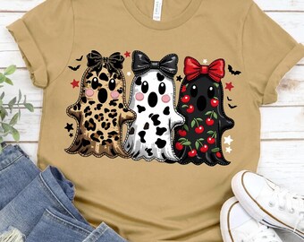 May include: A tan t-shirt featuring three cartoon ghosts with bows. The ghosts have different patterns: leopard print, black and white spots, and cherries. The design includes stars and bats.