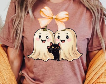 Puede incluir: Camiseta malva con un diseño de Halloween. El diseño incluye dos fantasmas sonrientes con lazos naranjas y un gato negro saludando. Los fantasmas son de color crema con mejillas rosadas.