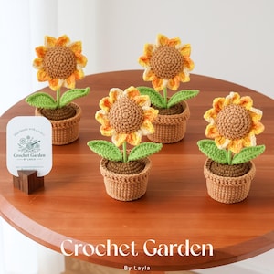 May include: Five crochet sunflowers in small brown pots sit on a wooden table. The sunflowers have yellow and orange petals, brown centers, and green leaves. A sign that says "Crochet Garden By Layla" is on the table.