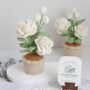 Peut inclure: Deux arrangements floraux blancs en crochet dans des pots marrons sur une surface blanche. Les fleurs sont dans un vase avec des feuilles vertes. Une carte de visite avec le texte "Handmade with Love" et "Crochet Garden by Layla" est au premier plan.