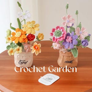 以下が含まれることがあります： 2つの鉢植えのクロシェ編みフラワーアレンジメント。鉢には「Enjoy」と「Best」の文字が書かれています。花は黄色、オレンジ、ピンク、パープル、ホワイトの色合いです。アレンジメントは、木製のテーブルの上に置かれ、「Crochet Garden」と書かれた名刺が置かれています。