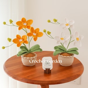 May include: Two potted crochet orchid plants, one with vibrant yellow flowers and the other with white flowers, each featuring green leaves and small buds on thin stems. The plants are in beige crocheted pots. A card reads "Crochet Garden".