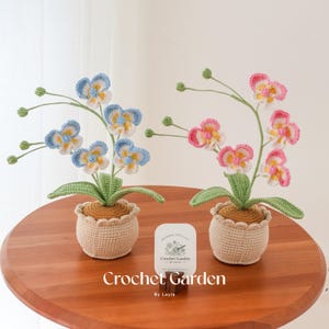 May include: Two crocheted orchid plants in pots. The orchid on the left has blue and white flowers, while the orchid on the right has pink and white flowers. Both plants are sitting on a wooden table with a sign that reads "Crochet Garden by Layla".