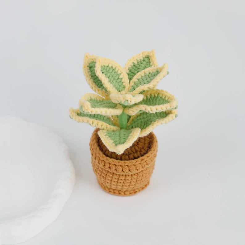 May include: A crocheted plant with green and yellow leaves sits in a brown crocheted pot. The plant has a detailed, textured appearance, and the pot has a woven design. The plant is a decorative item.