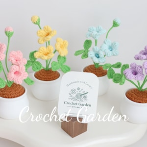 May include: Three crocheted flower arrangements in white pots. Each arrangement features different colored flowers: pink, yellow, blue, and purple. The pots are on a white surface with a sign that reads "Handmade with Love, Crochet Garden by Lily."