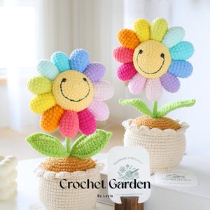 May include: Two crocheted rainbow flower pots with smiling faces. The pots are made of beige yarn and have a brown yarn base. The flowers have green leaves and are sitting on a white surface. The text "Crochet Garden by Layla" is on a white card.