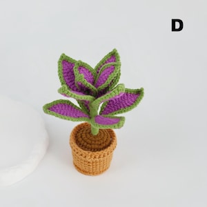 May include: A handmade crochet plant with purple and green leaves, set in a small brown pot. The plant has a detailed, textured appearance, showcasing the craftsmanship. The background is plain white, highlighting the vibrant colors of the plant.