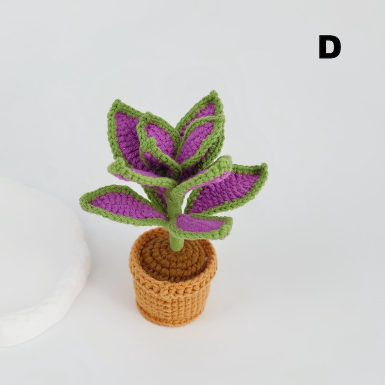 May include: A handmade crochet plant with purple and green leaves, set in a small brown pot. The plant has a detailed, textured appearance, showcasing the craftsmanship. The background is plain white, highlighting the vibrant colors of the plant.