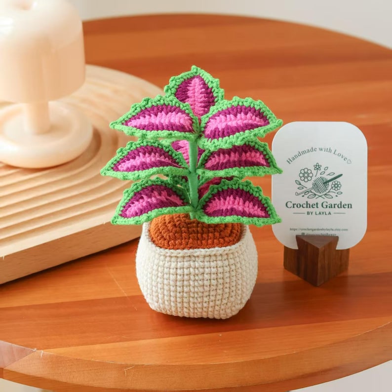 May include: A handmade crochet plant with pink and green leaves in a beige pot. The plant sits on a wooden table next to a card that reads "Handmade with Love" and "Crochet Garden - BY LAYLA".