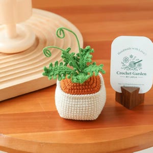 May include: A crocheted plant in a white pot with an orange top and green leaves. The plant has a tag that says "Handmade with Love" and "Crochet Garden by Layla."
