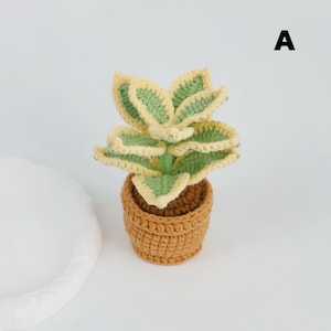 May include: A crocheted plant with green and yellow leaves sits in a brown crocheted pot. The plant has a stem and is labeled with the letter "A" in black. The plant is on a white surface.