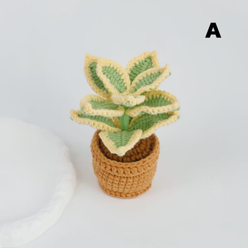 May include: A crocheted plant with green and yellow leaves sits in a brown crocheted pot. The plant has a stem and is labeled with the letter "A" in black. The plant is on a white surface.