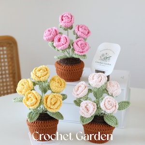 May include: Three crocheted flower arrangements in brown pots. The flowers are pink, yellow, and white. The pots are brown and have a crocheted texture. The flowers are arranged in a bouquet style. The image includes the text "Handmade with Love" and "Crochet Garden by Layla".