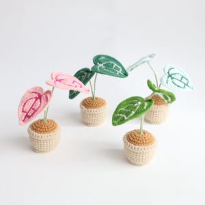 May include: A collection of handmade crochet plants in small pots. The leaves are various colors, including pink, green, and blue. The pots are a light beige color, and the leaves have detailed stitching.