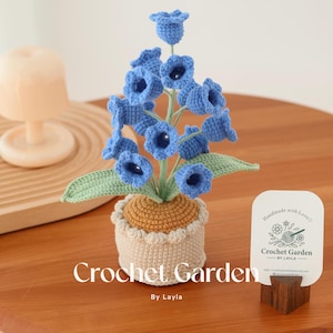 May include: Crocheted bluebell flowers in a cream and brown pot with green leaves. The pot is sitting on a wooden table. The image includes a business card that says "Crochet Garden by Layla".
