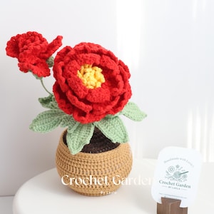 Puede incluir: Un arreglo de flores de peonía rojas de ganchillo en una maceta marrón con hojas verdes. El arreglo está sobre una superficie blanca con una tarjeta de visita que dice "Handmade with love, Crochet Garden by Layla".