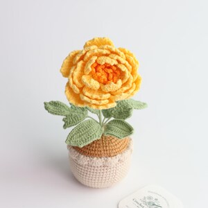 May include: A handcrafted crochet flower with layered yellow and orange petals, featuring sage green leaves on a stem. The flower is potted in a two-toned beige and tan crocheted container with a textured, rounded base and a scalloped top edge.