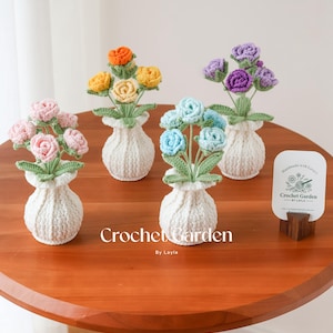 May include: Four crocheted flower arrangements in white vases on a wooden table. The flowers are pink, orange, blue, and purple. A sign reads "Handmade with Love, Crochet Garden by Layla".