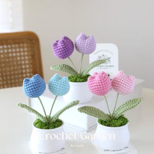 May include: Three crocheted tulip flower arrangements in white pots. The tulips are in shades of purple, blue, and pink. The pots are decorated with green moss. The label on the pot reads "Crochet Garden by Layla".