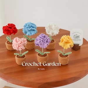 Könnte beinhalten: Eine Sammlung von sieben gehäkelten Nelkenblüten in verschiedenen Farben, jede in einem braunen gehäkelten Topf. Die Blumen sind auf einem Holztisch mit einem Schild angeordnet, auf dem "Handmade with Love Crochet Garden By Layla" steht.